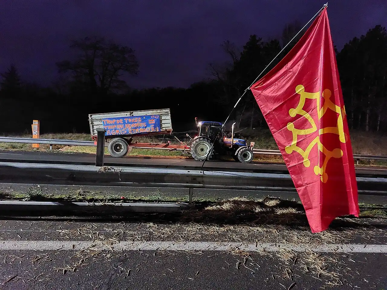 De nuit, un tracteur et sa remorque portant une banderole « Trop de taxes, trop de normes, agris étranglés » sur une autoroute ; au premier plan, un drapeau occitan rouge et jaune.
