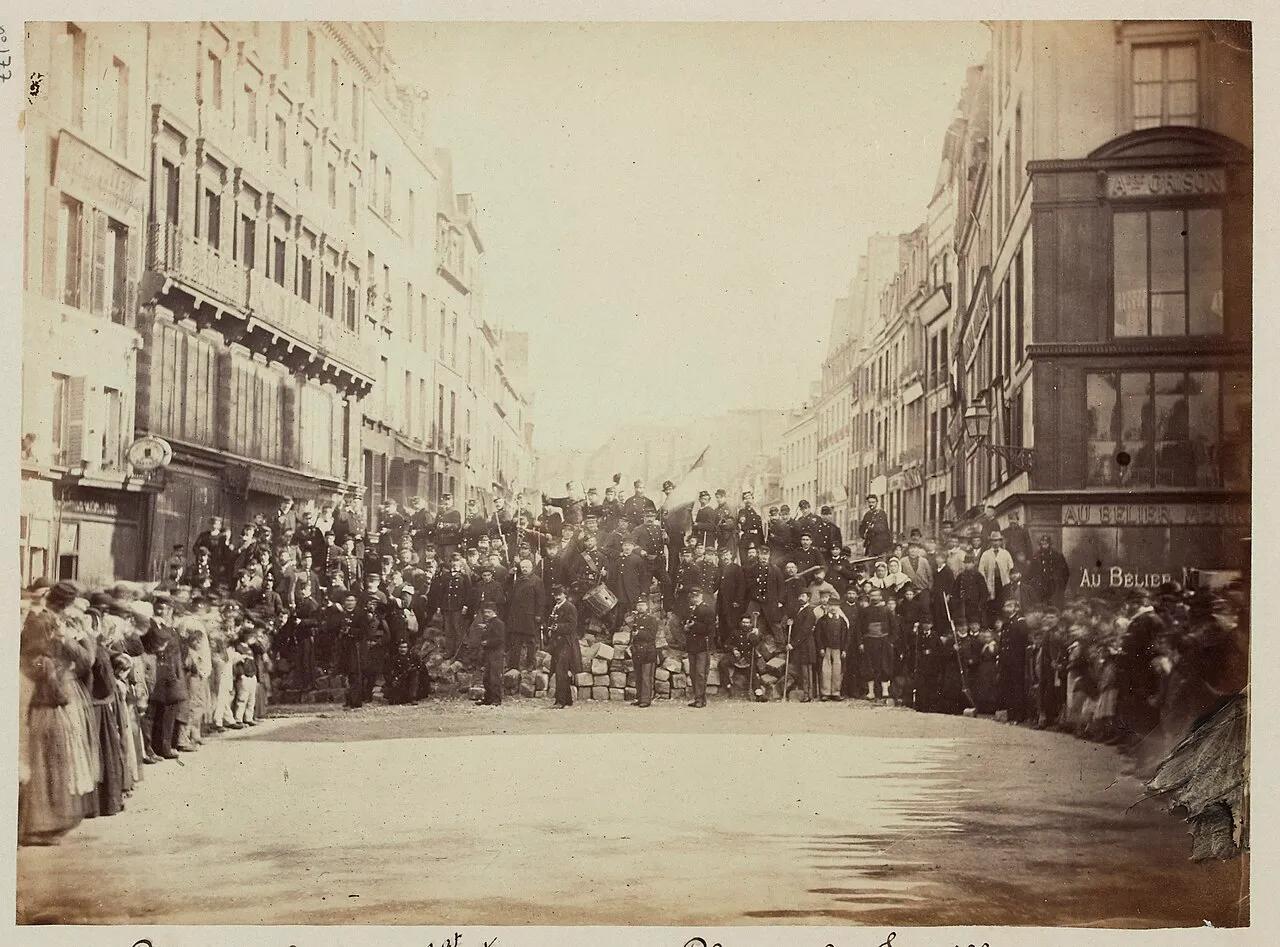 Photographie sépia d'une rue bordée d'immeubles. Au centre, une barricade basse de pavés sur laquelle posent des dizaines d'hommes en uniforme, dont un tambour ; des passants, femmes et enfants, se tiennent sur les côtés.