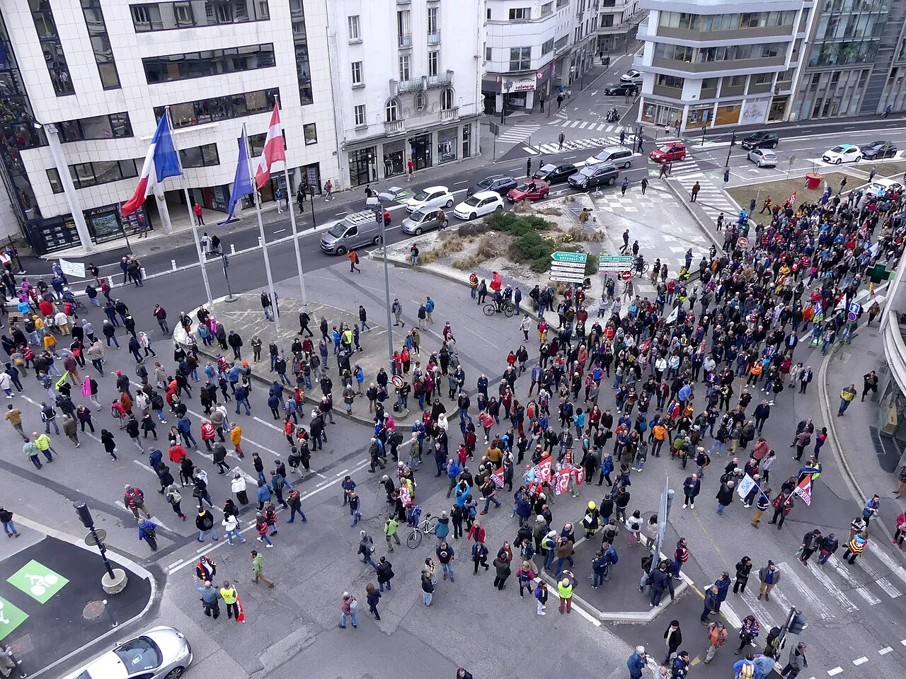 Vue plongeante sur une place et un carrefour remplis de manifestants, avec drapeaux et pancartes, entre des immeubles de ville.