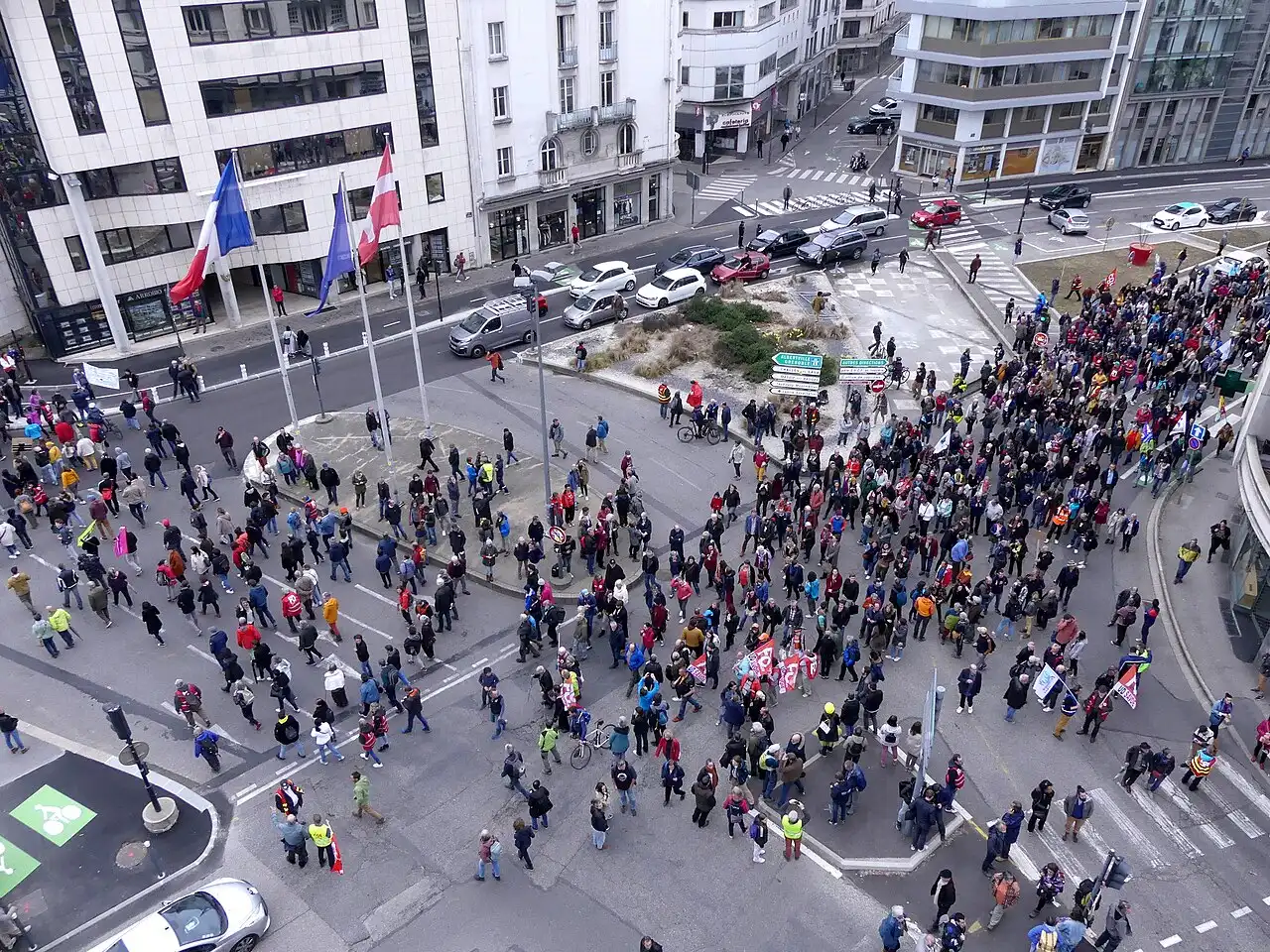 Vue plongeante sur une place et un carrefour remplis de manifestants, avec drapeaux et pancartes, entre des immeubles de ville.