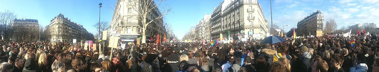 Vue panoramique d'une foule dense sur une avenue parisienne ensoleillée, avec des pancartes et des drapeaux arc-en-ciel.