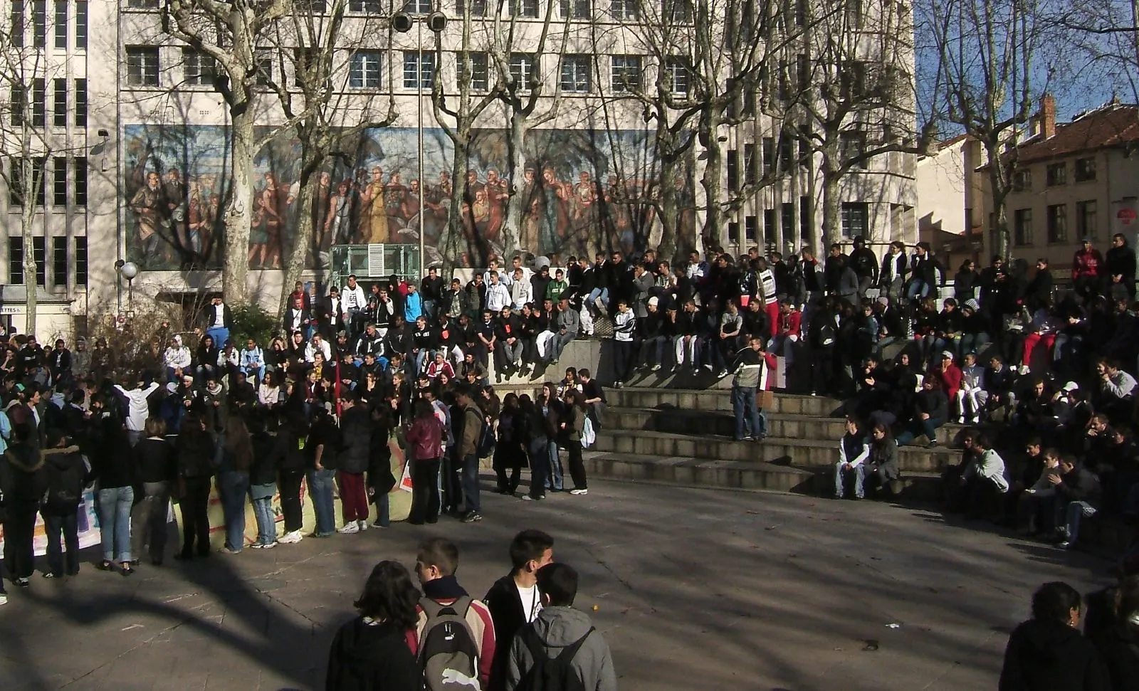 Foule de jeunes rassemblés sur les marches et la place devant un bâtiment orné d'une grande fresque, sous des platanes sans feuilles.