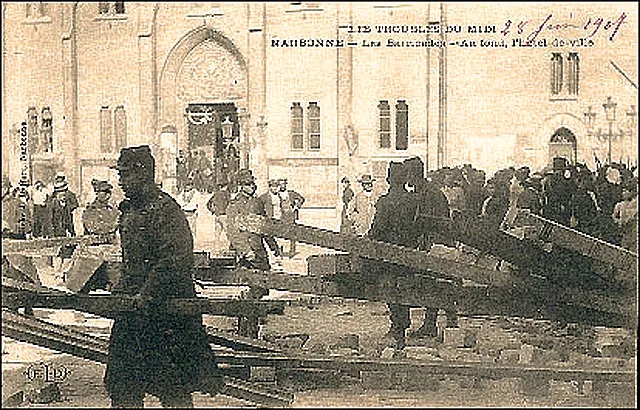 Carte postale sépia : des poutres et des pavés renversés barrent une place ; un soldat en capote au premier plan, des groupes d'hommes devant un bâtiment à grande porte en arc.