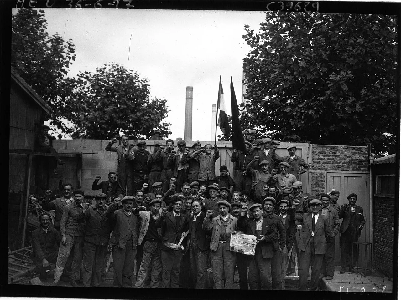 Une soixantaine d'ouvriers en casquette posent poing levé dans une cour d'usine, devant un drapeau tricolore et un drapeau sombre ; l'un tient un exemplaire de L'Humanité.