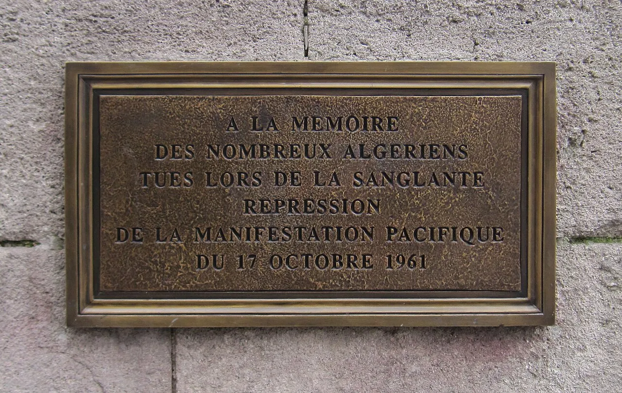 Plaque de bronze rectangulaire fixée sur un mur de pierre, portant une inscription en lettres capitales.