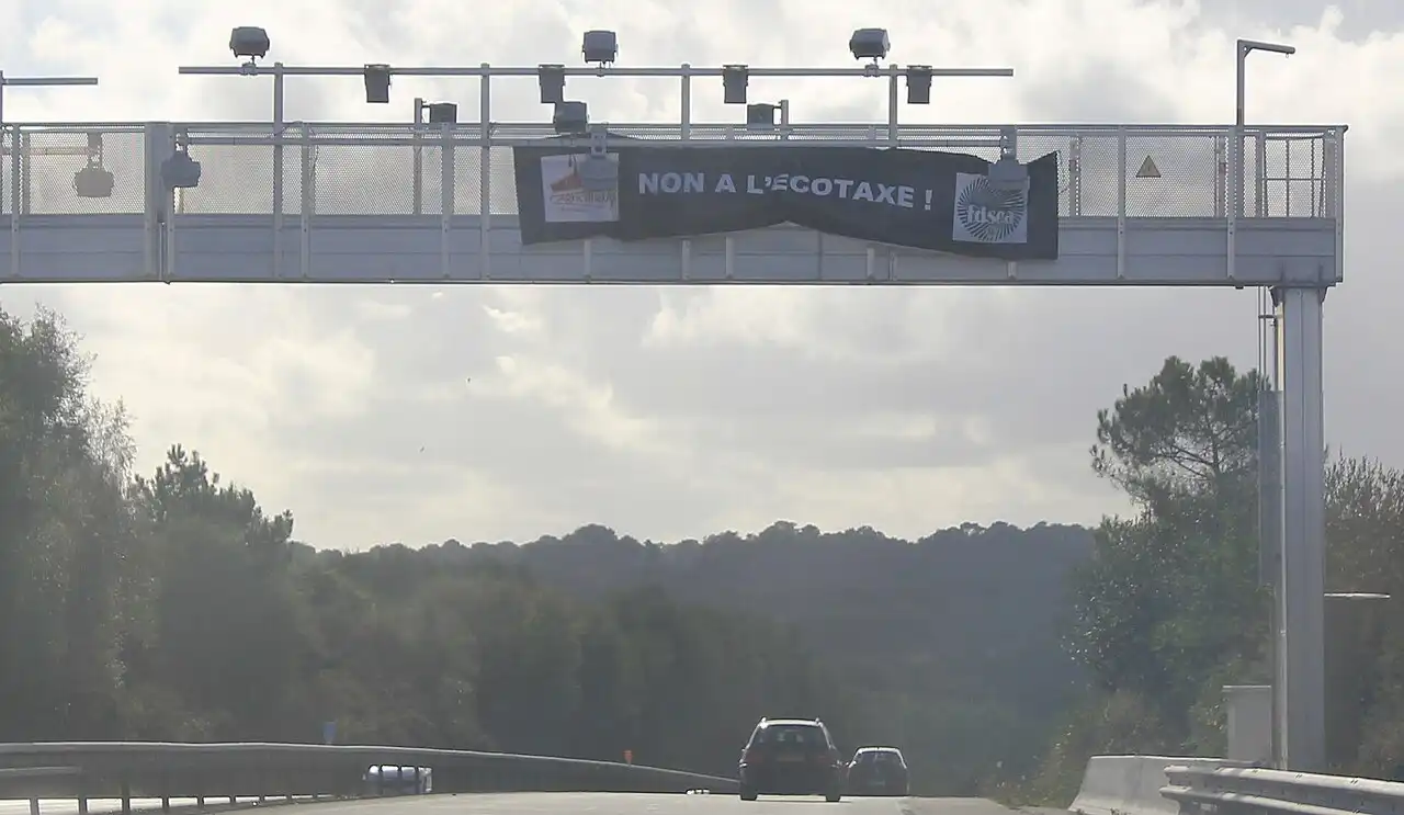 Portique métallique au-dessus d'une voie rapide, équipé de caméras, portant une banderole noire « Non à l'écotaxe ! » ; deux voitures passent dessous.