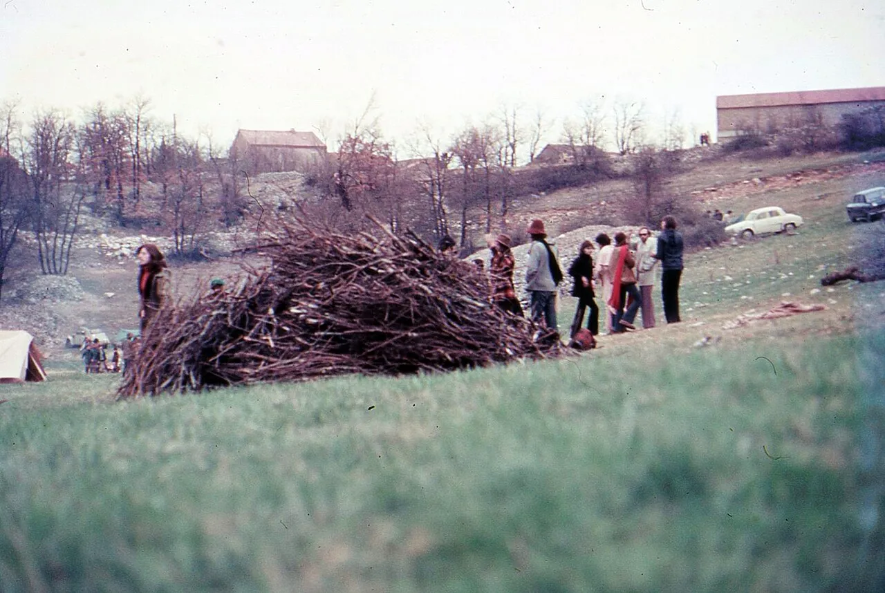 Photographie en couleur d'un groupe de personnes debout dans un pré à côté d'un grand tas de branchages, avec des fermes et des voitures en arrière-plan.