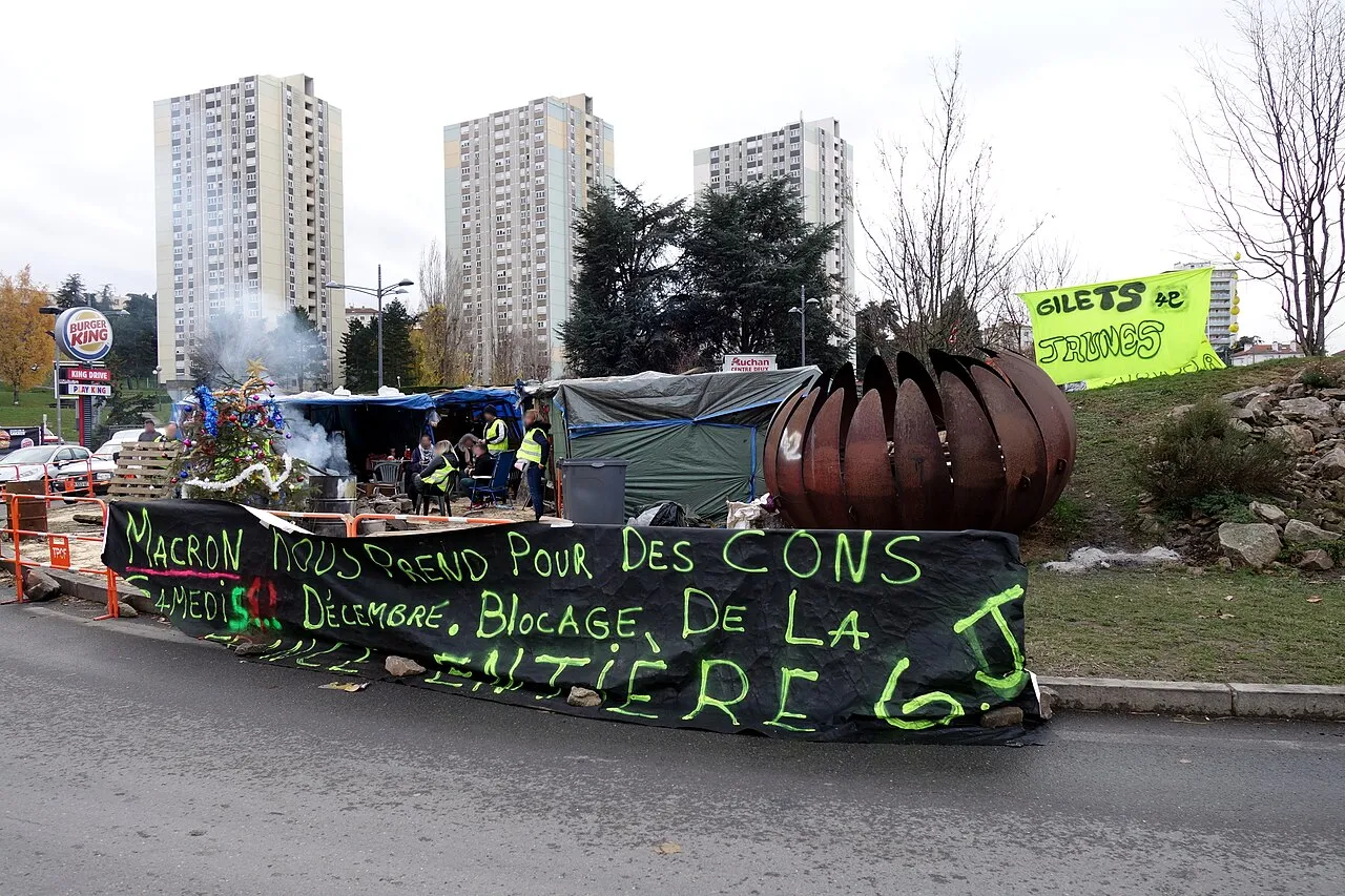 Campement de tentes et de bâches sur un rond-point, avec un sapin de Noël décoré, une grande banderole noire écrite en vert fluo et des immeubles en arrière-plan.